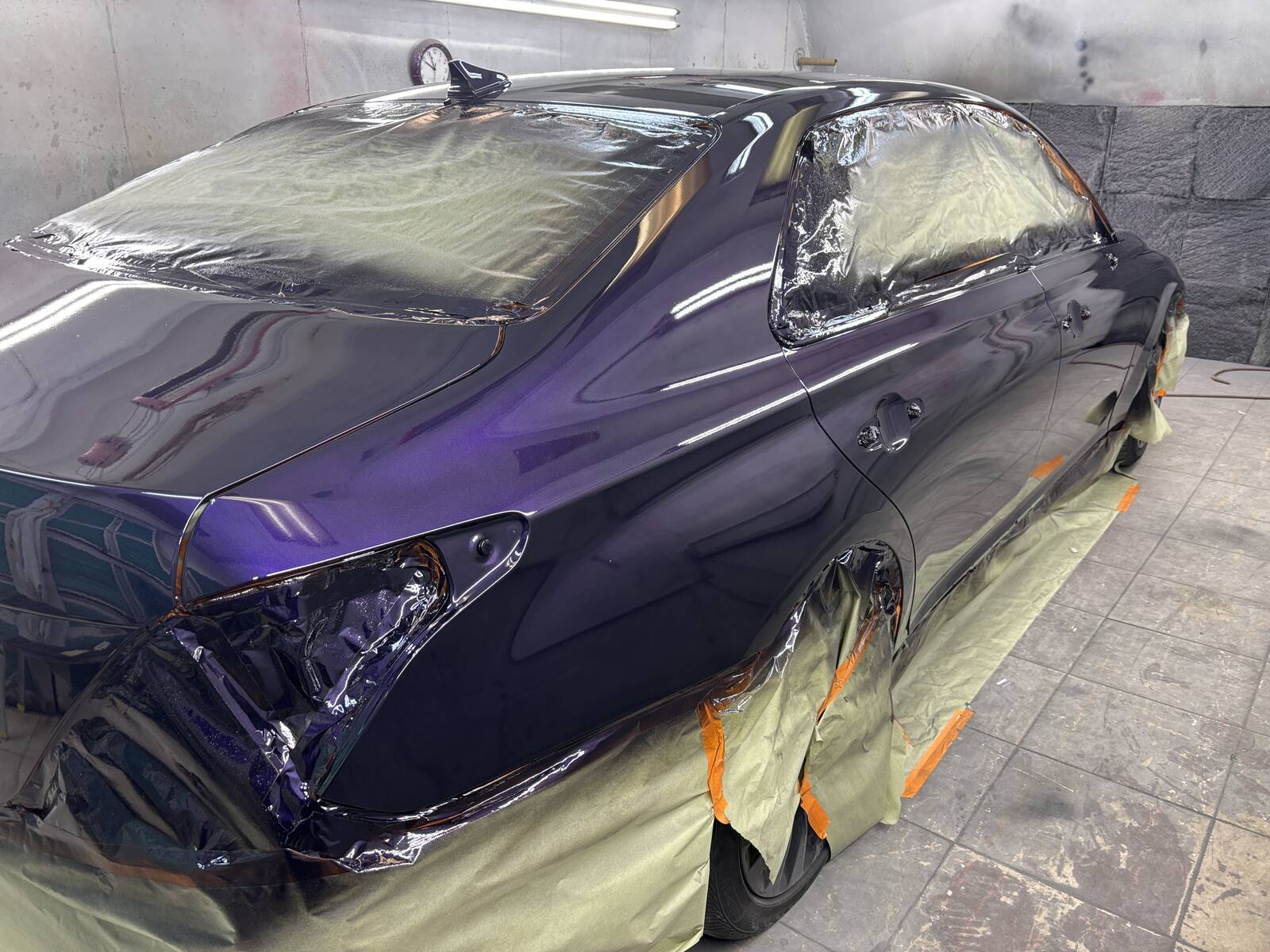 A dark purple sedan masked off in the paint booth with fresh, wet paint on the side.